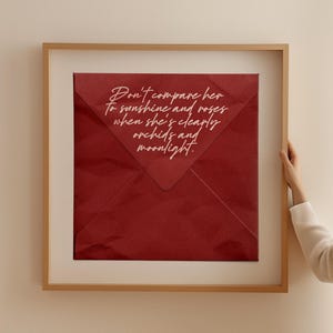 以下が含まれることがあります： 赤い封筒に「彼女を太陽やバラに例えないで。彼女は明らかに蘭と月光なのだから。」という白い文字が書かれた額装されたプリントです。