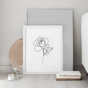Puede incluir: Un dibujo en blanco y negro de una rosa en un marco blanco. El marco est&aacute; sobre un suelo de madera con una cesta de mimbre y un jarr&oacute;n de vidrio.