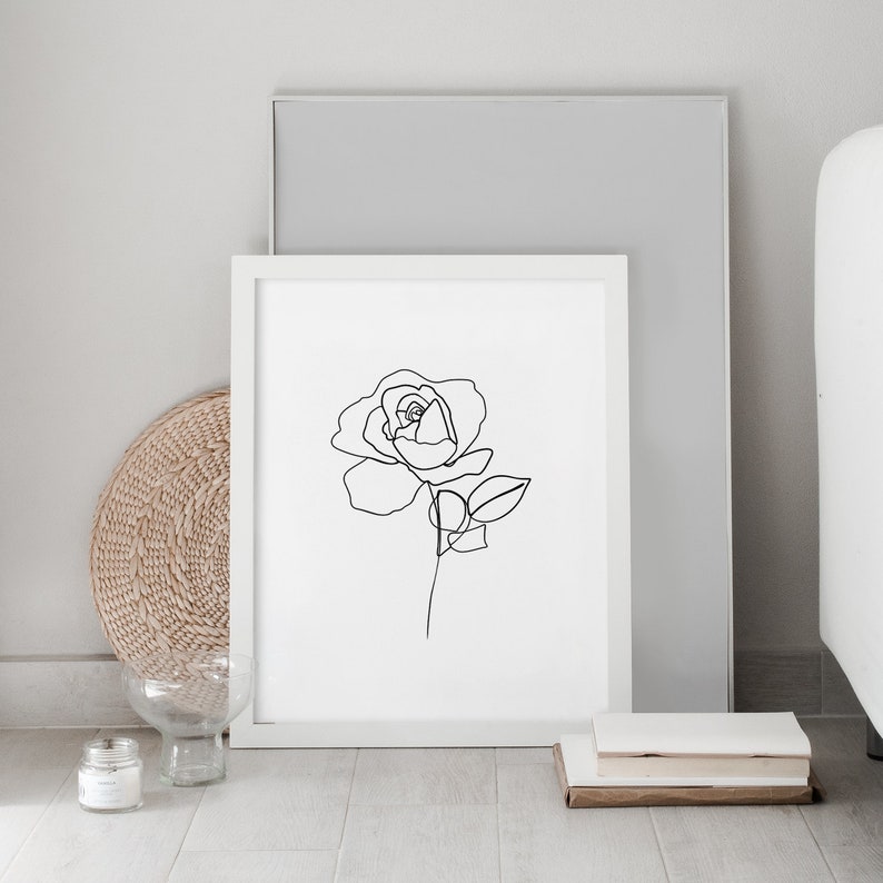 Puede incluir: Un dibujo en blanco y negro de una rosa en un marco blanco. El marco est&aacute; sobre un suelo de madera con una cesta de mimbre y un jarr&oacute;n de vidrio.