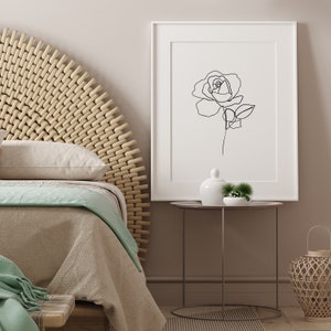 Puede incluir: Un dibujo en blanco y negro de una rosa en un marco blanco colgado en una pared sobre una mesita de noche con un jarr&oacute;n de cer&aacute;mica blanco y una peque&ntilde;a planta verde.