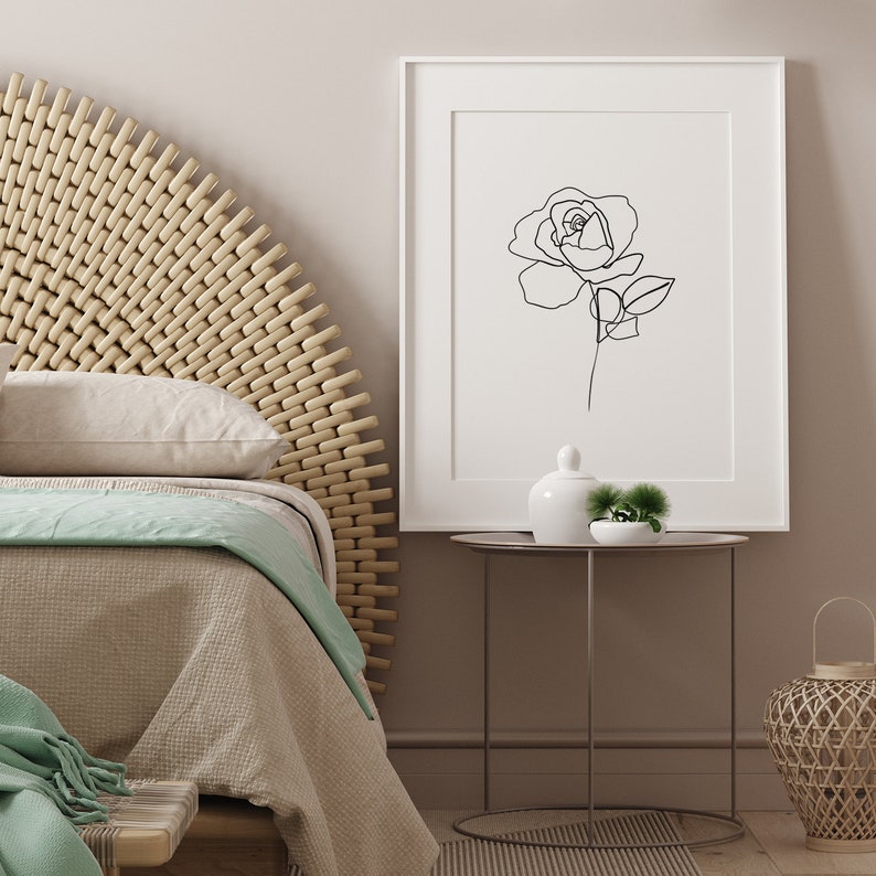 Puede incluir: Un dibujo en blanco y negro de una rosa en un marco blanco colgado en una pared sobre una mesita de noche con un jarr&oacute;n de cer&aacute;mica blanco y una peque&ntilde;a planta verde.