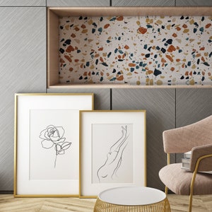 Puede incluir: Dos dibujos en blanco y negro enmarcados en una pared. El dibujo de la izquierda es de una sola rosa. El dibujo de la derecha es de una persona con los brazos levantados por encima de la cabeza. La pared es de color gris claro con un patr&oacute;n de veta de madera.