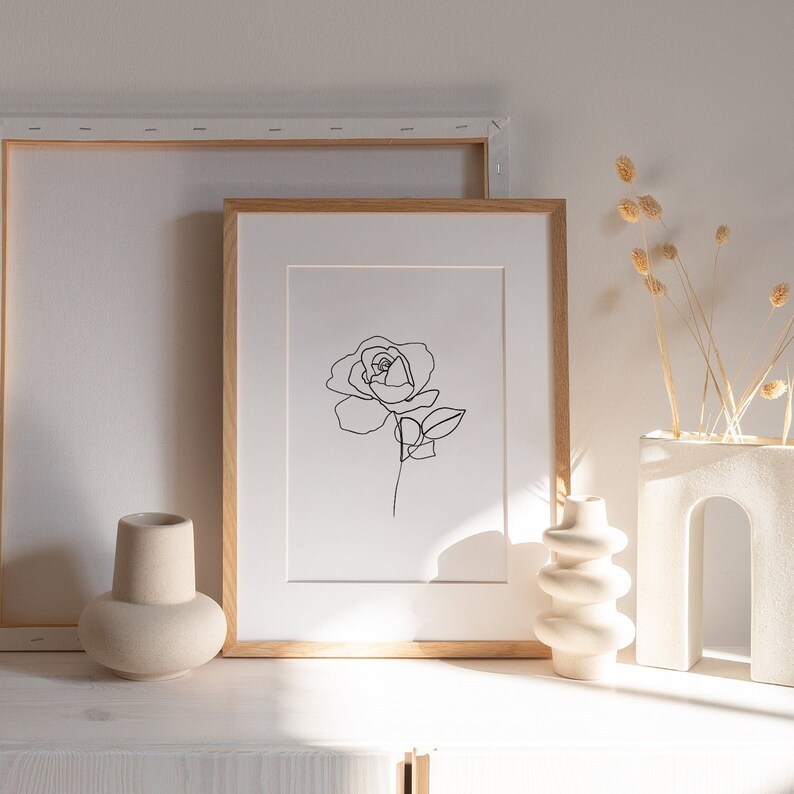Puede incluir: Un dibujo en blanco y negro de una sola rosa en un estilo minimalista, enmarcado. El marco es de madera clara y el dibujo est&aacute; sobre papel blanco.