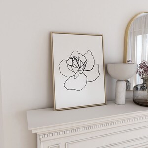 Puede incluir: Un dibujo en blanco y negro de una rosa en un estilo simple y minimalista. El dibujo est&aacute; enmarcado en un marco de madera clara y se muestra en una repisa blanca.