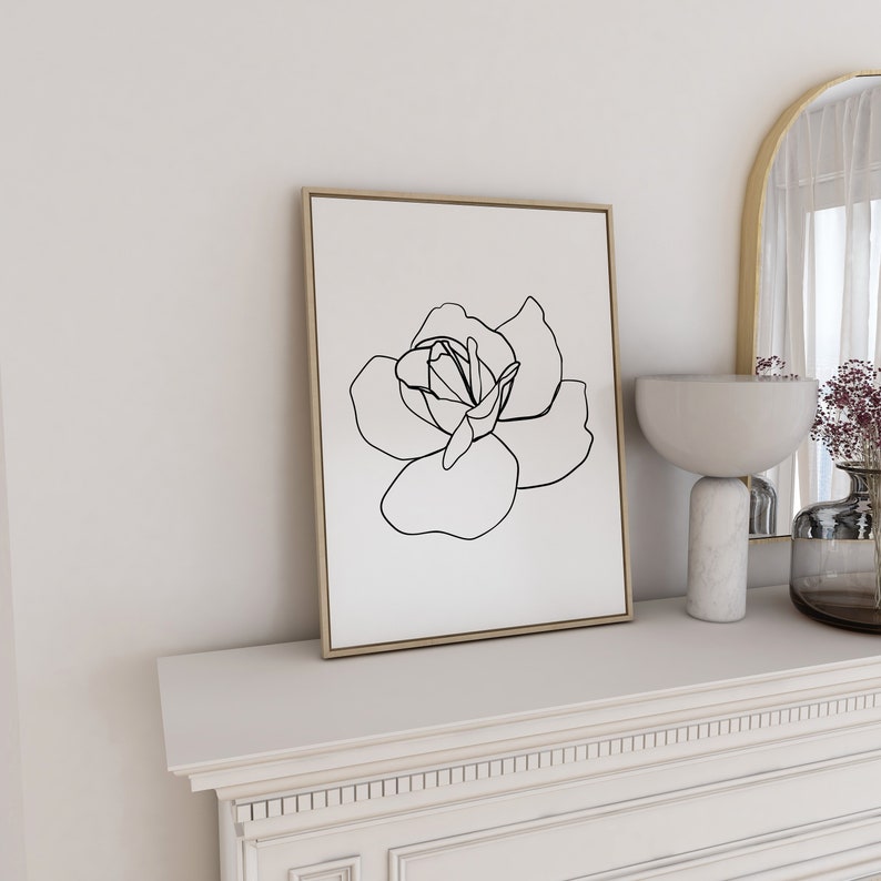 Puede incluir: Un dibujo en blanco y negro de una rosa en un estilo simple y minimalista. El dibujo est&aacute; enmarcado en un marco de madera clara y se muestra en una repisa blanca.