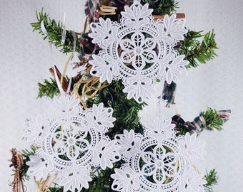 May include: Three white lace snowflake ornaments hanging on a green Christmas tree. The snowflakes are delicate and intricate, with a floral design in the center.