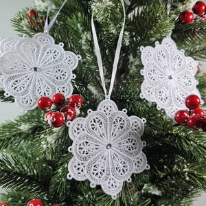 May include: Three white snowflake decorations with intricate lace designs hang on a green Christmas tree with red berries.