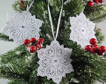 May include: Three white snowflake ornaments with intricate lace designs hang on a green Christmas tree with red berries.