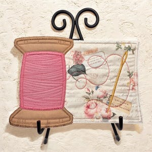 May include: A fabric wall hanging featuring a tan spool of pink thread on the left and a rectangular panel on the right with a floral print, a gold needle, and the text "LIVE LIFE IN FULL BLOOM" and "WITH ALL OF YOUR HEART" on a cream background.
