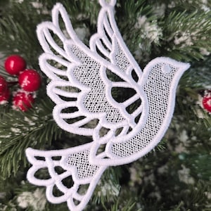 May include: A white, lace dove Christmas tree ornament with intricate wing and tail detailing, suspended by a white ribbon. The dove is set against a backdrop of green fir branches and red berries, creating a festive holiday scene.