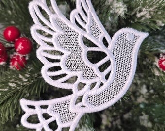 May include: A white, lace dove Christmas tree ornament with intricate wing and tail detailing, suspended by a white ribbon. The dove is set against a backdrop of green fir branches and red berries, creating a festive holiday scene.