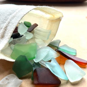 May include: A white fabric bag spills out a variety of sea glass pieces in shades of green, blue, and brown. The glass pieces are smooth and have been tumbled by the sea.