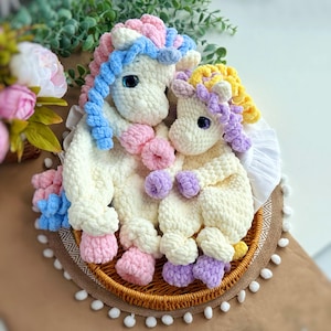 May include: Two handmade crochet unicorns in cream, with blue, pink, and yellow manes and tails. The unicorns are nestled together on a round woven tray with a decorative trim. The background includes greenery and flowers.