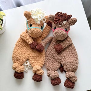 May include: Two crocheted plush horse toys. One is brown with a pink nose and the other is tan with a blue nose. Both have white yarn for manes and tails.
