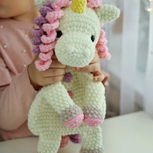 Può includere: Un peluche di unicorno bianco all'uncinetto con criniera e coda in lana rosa e viola. L'unicorno ha un corno giallo e occhi neri.