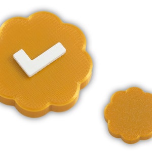 May include: Two gold-colored, flower-shaped objects on a white background. The larger object has a white checkmark in the center. The smaller object is a solid gold color. The objects appear to be 3D printed.