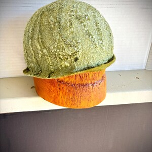 May include: A green velvet cloche hat with a wide brim, sitting on a wooden hat form.