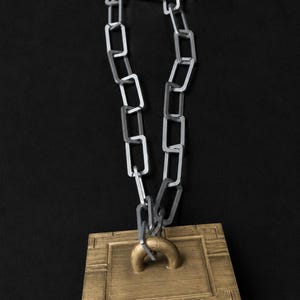 May include: A bronze-colored, square-shaped pendant with a curved handle, connected to a silver chain. The pendant has a textured surface and is suspended against a black background. The chain is made of rectangular links.