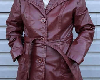 Può includere: Una giacca di pelle bordeaux con vita a cintura e un colletto con un dettaglio decorativo. La giacca ha una chiusura con bottoni e due tasche.