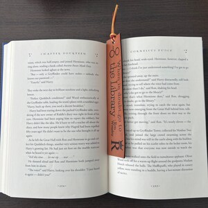 Close up of dark brown leather bookmark laser engraved with wizard design + quote with suede cord tassel in red and gold laying inside open book to show size