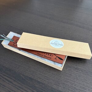 Dark brown leather bookmark laser engraved with wizard design and suede cord tassel shown inside a kraft paper gift box with shop logo