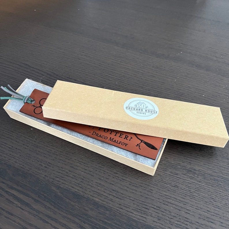 Dark brown leather bookmark laser engraved with wizard design and suede cord tassel shown inside a kraft paper gift box with shop logo