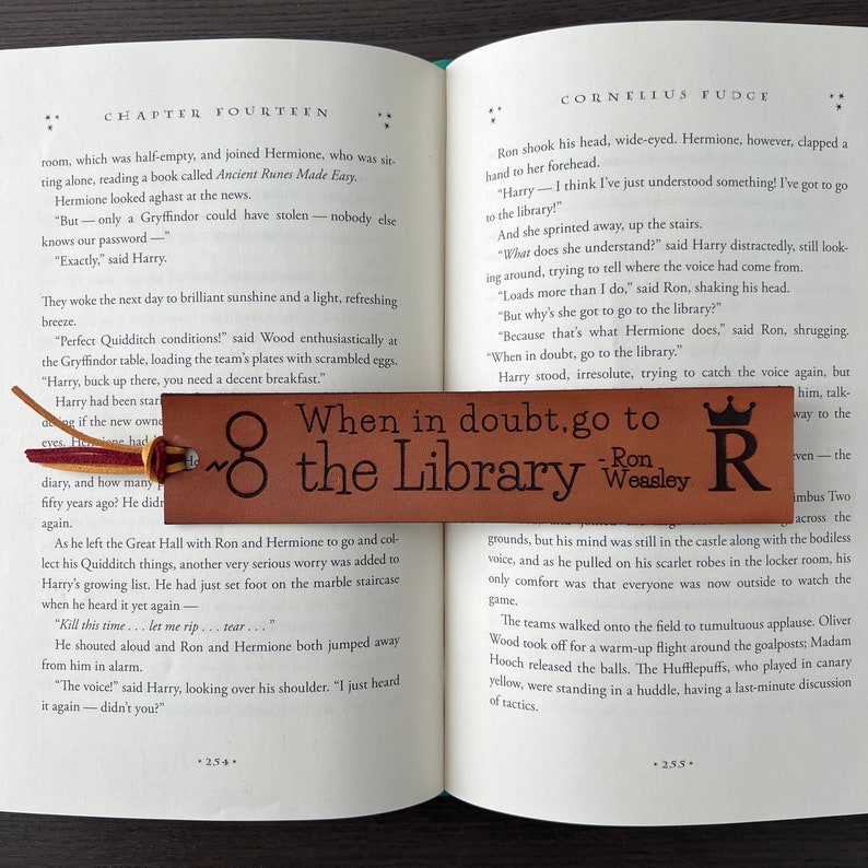 Close up of dark brown leather bookmark laser engraved with wizard design + quote with suede cord tassel in red and gold laying inside open book to show size