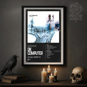 May include: Framed poster for the Radiohead album "OK Computer" with a black frame. The poster features a blue and white graphic with the album title and tracklist. A black crow statue, books, candles, and a skull are on a dark wooden surface.