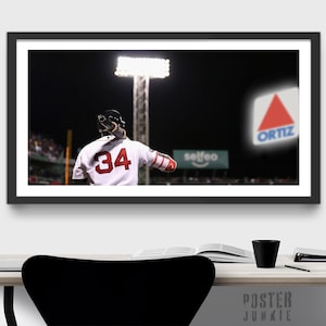May include: A baseball player wearing a white jersey with the number 34 in red, stands on a baseball field at night. The player is facing away from the camera and holding a bat. The player is standing in front of a large sign that says "Ortiz" in white letters on a red triangle.