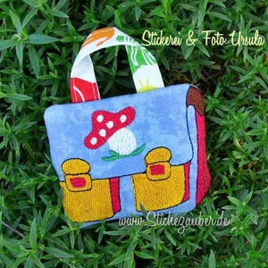 May include: A small blue fabric backpack with a red mushroom design and yellow buckles. The backpack has a red and white striped handle and the text "www.Stichezauber.de" is printed on the bottom.