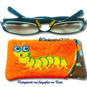May include: Orange fabric glasses case with a green and yellow embroidered caterpillar wearing blue glasses. The text "Probegestickt und fotografiert von Britta" is at the bottom of the case.