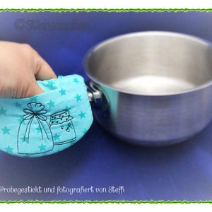 Könnte beinhalten: Ein türkisfarbener Topflappen mit Sternenmuster und gestickten Bildern eines Glases und einer Tasche. Der Topflappen wird verwendet, um einen silbernen Topf zu halten.