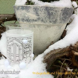 Könnte beinhalten: Ein weißer Keramiktopf mit grauer Glasur steht im Schnee. Im Vordergrund steht ein Glas mit einer schwarz-weißen Illustration eines Schneemanns vor einem Haus. Das Glas ist teilweise mit Schnee bedeckt.