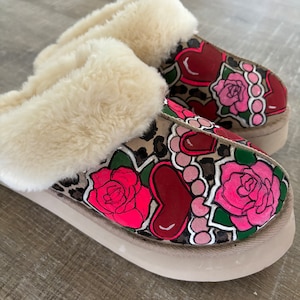 May include: A pair of fuzzy slippers with a leopard print base and a hand-painted design featuring pink roses, red hearts, and green leaves.
