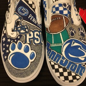 May include: A pair of white canvas slip-on shoes with a custom design featuring the Penn State Nittany Lions logo, a football, and other sports-related imagery. The shoes are painted in shades of blue, green, brown, and black.