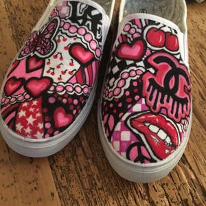 May include: A pair of white canvas slip-on shoes with a colorful, hand-painted design. The design features hearts, cherries, stars, and a Chanel logo. The shoes are a fun and unique way to express your personality.