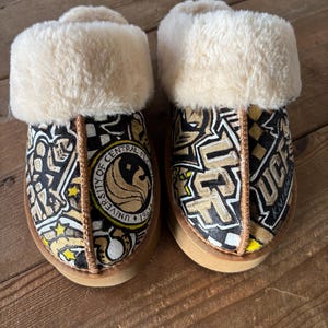May include: A pair of brown and black slippers with a white faux fur lining. The slippers feature a black and white checkered pattern with a gold and black University of Central Florida logo.