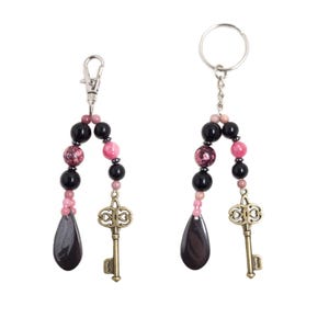 Puede incluir: Dos llaveros con hebras de cuentas en negro, rosa y morado brillante. Cada llavero tiene un dije de llave de color bronce y una cuenta en forma de lágrima. Un llavero tiene un cierre, el otro un llavero.