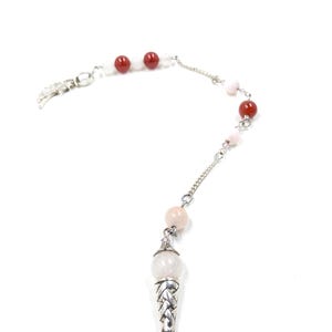 May include: A silver-toned pendulum with a light pink crystal at the bottom and a chain with red and white beads. The pendulum has a decorative braided design and a small pink crystal at the tip.