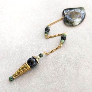 May include: A pendulum featuring a polished heart-shaped stone in shades of gray and white, connected by a gold-toned chain to a decorative gold-toned metal cone. The chain is accented with small black and green beads throughout its length.