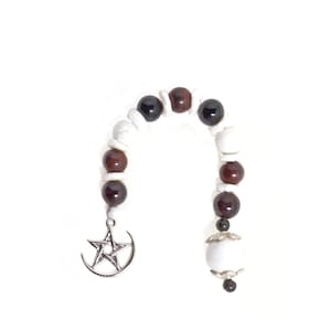Puede incluir: Una pulsera de cuentas con cuentas blancas, rojo oscuro y negras. Un dije plateado con una estrella y una luna creciente cuelga de la pulsera. La pulsera está sobre un fondo blanco.
