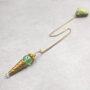 May include: A gold-toned metal pendulum featuring a conical base with intricate embossed patterns, a translucent light green spherical bead, and small clear crystal accents. The pendulum is attached to a fine gold-toned chain with a green stone.