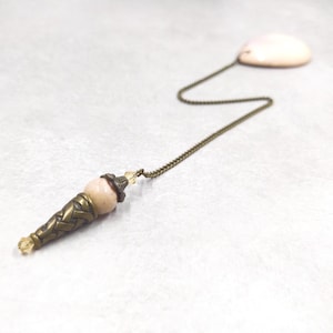 May include: A pendulum featuring a smooth, pale pink stone bead topped with an ornate, antique brass-toned metal cone and a small clear crystal bead. The piece is attached to a thin, antique brass-toned metal chain connected to a flat, pale pink stone.