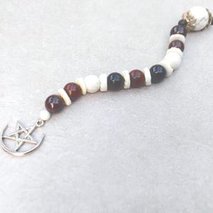 May include: A beaded charm featuring a silver-toned pentagram and crescent moon pendant. The strand consists of alternating round beads in white marble, deep red, and black, separated by small flat white spacers, ending with a decorative metal cap.