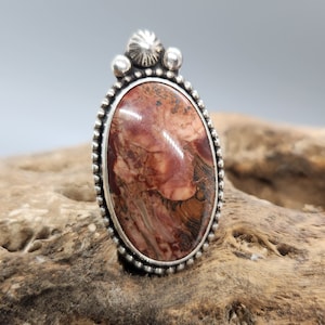 May include: A silver ring with a red, brown, and white stone set in an oval bezel. The ring has a beaded edge and a decorative top.