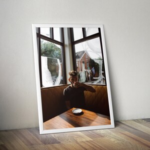 May include: A man sits at a table in a cafe, looking out the window. He is wearing a dark shirt and has a cup of coffee in his hand. The cafe has large windows and a wooden table.