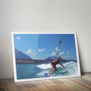 May include: A person wakeboarding on a lake with mountains in the background. The person is wearing a red shirt and blue shorts. The water is blue and the sky is clear.