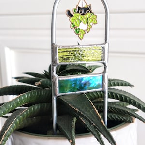 May include: A metal plant stake with three stained glass panels in shades of green and blue. A black cat with green leaves hangs from the top of the stake.