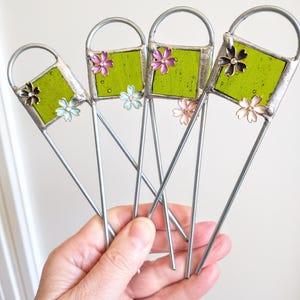 May include: Five green glass squares with metal frames and colourful flower accents. The squares are attached to long, thin metal rods.