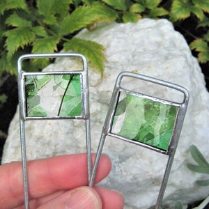 May include: Two silver metal garden markers with green stained glass squares. The markers are held in a hand.